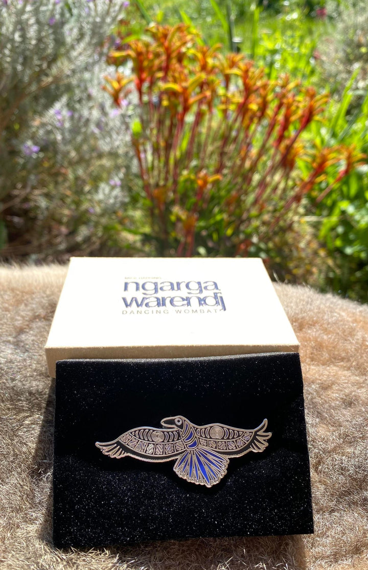 Add a finishing touch to any outfit, with this Lapel Pin featuring a Waang the Crow design by Ngarga Warendj Dancing Wombat Measures 26.5mm x 60mm  Presented in a stylish box with magnetic closure  Made with metal with enamel inlay Includes an information card on the design and artist