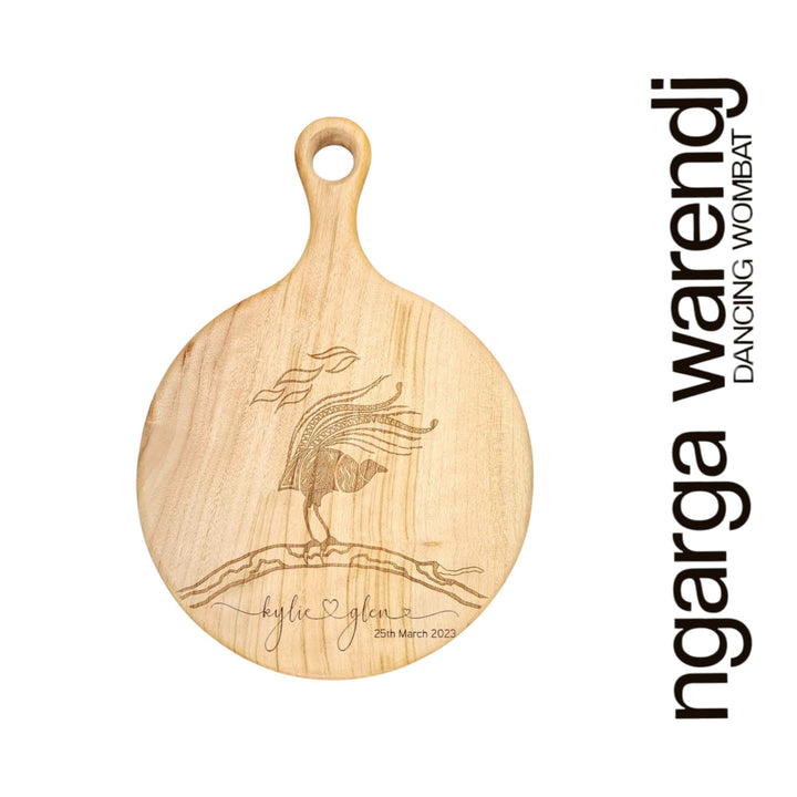 Wooden cutting board with laser engraving of a lyrebird design and 'ngarga warendj' branding on a white background.