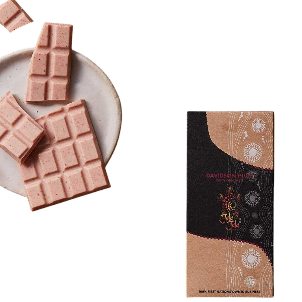 Pink chocolate bars shown broken on a light ceramic plate alongside the product packaging box, which features a black and tan Aboriginal-inspired pattern with the brand name Davidson Plum White Chocolate.