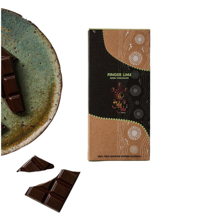 A bar of Finger Lime dark chocolate shown in its packaging alongside two broken pieces of the chocolate, with a cropped ceramic plate on the left edge.