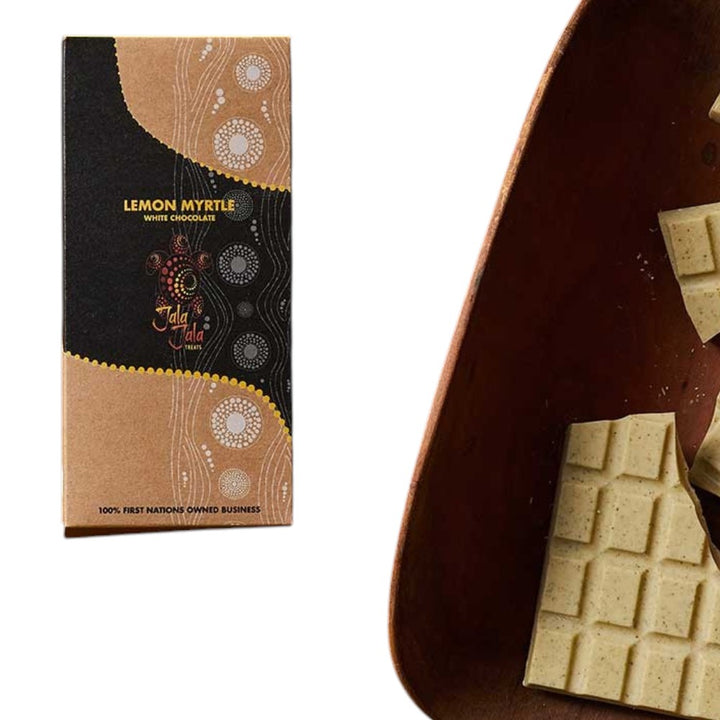 A chocolate bar package labeled 'LEMON MYRTE WHITE CHOCOLATE' stands beside a close-up of a dark brown chocolate bar broken into pale yellow minty pieces on a white background.