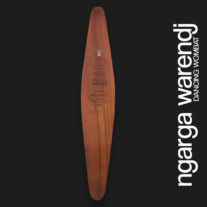 A long, narrow wooden board or plaque with a warm brown wood grain and engraved text, centered on a black background. Large white vertical text along the right side reads 'ngarga warendj DANCING WOMBAT'.