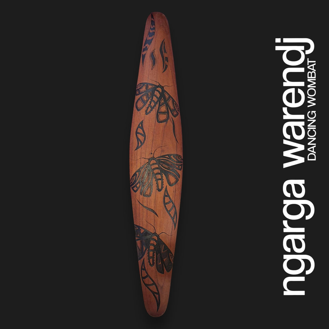 A tall, narrow wooden wall-hanging or decorative plaque with a warm brown wood tone and black bogong moths motifs, shown against a dark charcoal background. Large white vertical text on the right reads 'ngarga warendj' with smaller text 'Dancing Wombat'.