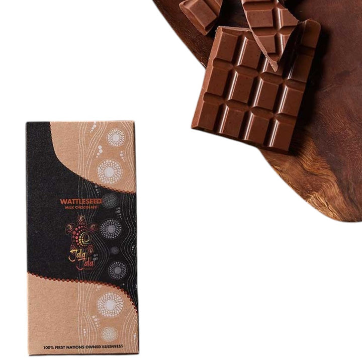 A chocolate bar package standing upright on the left, next to a broken chocolate bar on a dark wooden surface against a white background. The package is tan and black with circular white patterns and the brand name Wattleseed.